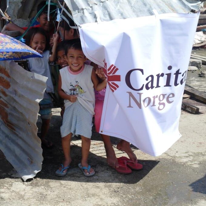 Children smiling under a white flag of Caritas Norge